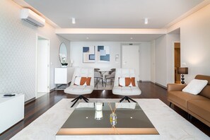 A modern living room with a glass coffee table, two white armchairs with orange cushions, a brown sofa, and a white cabinet.
