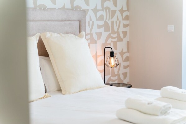 A neatly made bed with white linens and pillows, a bedside table with a geometric light fixture, and a patterned wall in the background.