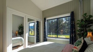 A bedroom with a large bed, a window with curtains, a plant, and a view of the outdoors.