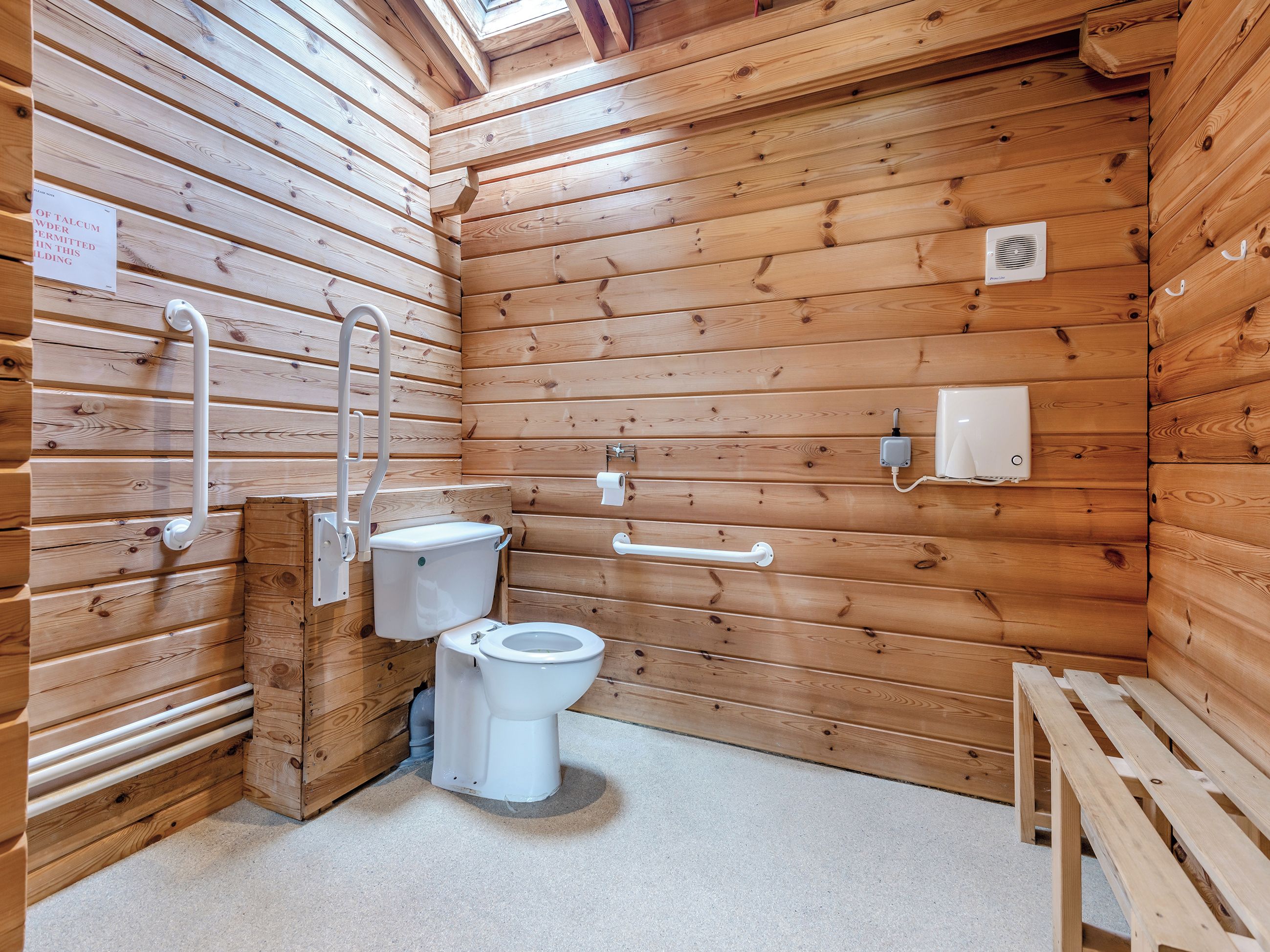 Une salle de bain rustique, aménagée dans une cabane en bois, avec des toilettes, des barres d’appui et un banc.