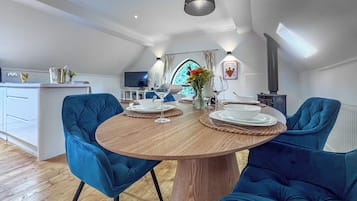 Un espace repas moderne avec une table ronde en bois, deux chaises bleues à revêtement rembourré, un téléviseur en arrière-plan et une cuisine visible à travers une fenêtre ouverte.