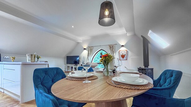 A modern dining area with a round wooden table, two blue upholstered chairs, a television in the background, and a kitchen visible through an open window.