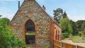 Un bâtiment en pierre avec un toit triangulaire, une grande fenêtre en arc de cercle et une clôture en bois à l’avant.