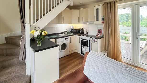 A compact kitchen with a washing machine, a microwave, and a small oven. There is a sofa and a window with curtains.