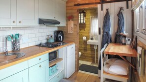 A compact kitchen with white cabinets, a stove, and a sink. There is a small dining area with a chair and a table. - Ty'r Bugail Shepherds Hut (Tal Y Bont, near Barmouth)