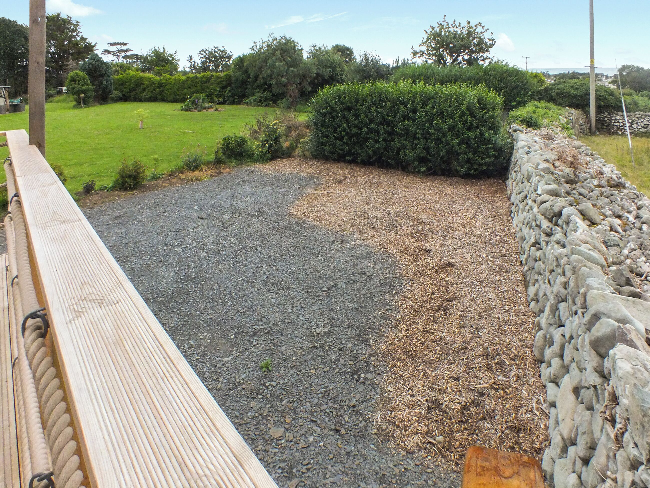 A wooden deck with a stone wall and a gravel area with a hedge.