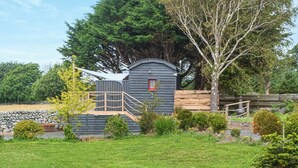 A small cabin with a wooden deck and a red window, surrounded by a stone wall and lush greenery. - Ty'r Bugail Shepherds Hut (Tal Y Bont, near Barmouth)