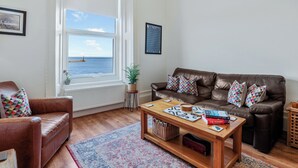 A living room with a brown leather sofa, a wooden coffee table with books, a window with a view of the sea, and a framed picture on the wall. - 3 bedroom accommodation in Roker, near Sunderland (Roker, near Sunderland)
