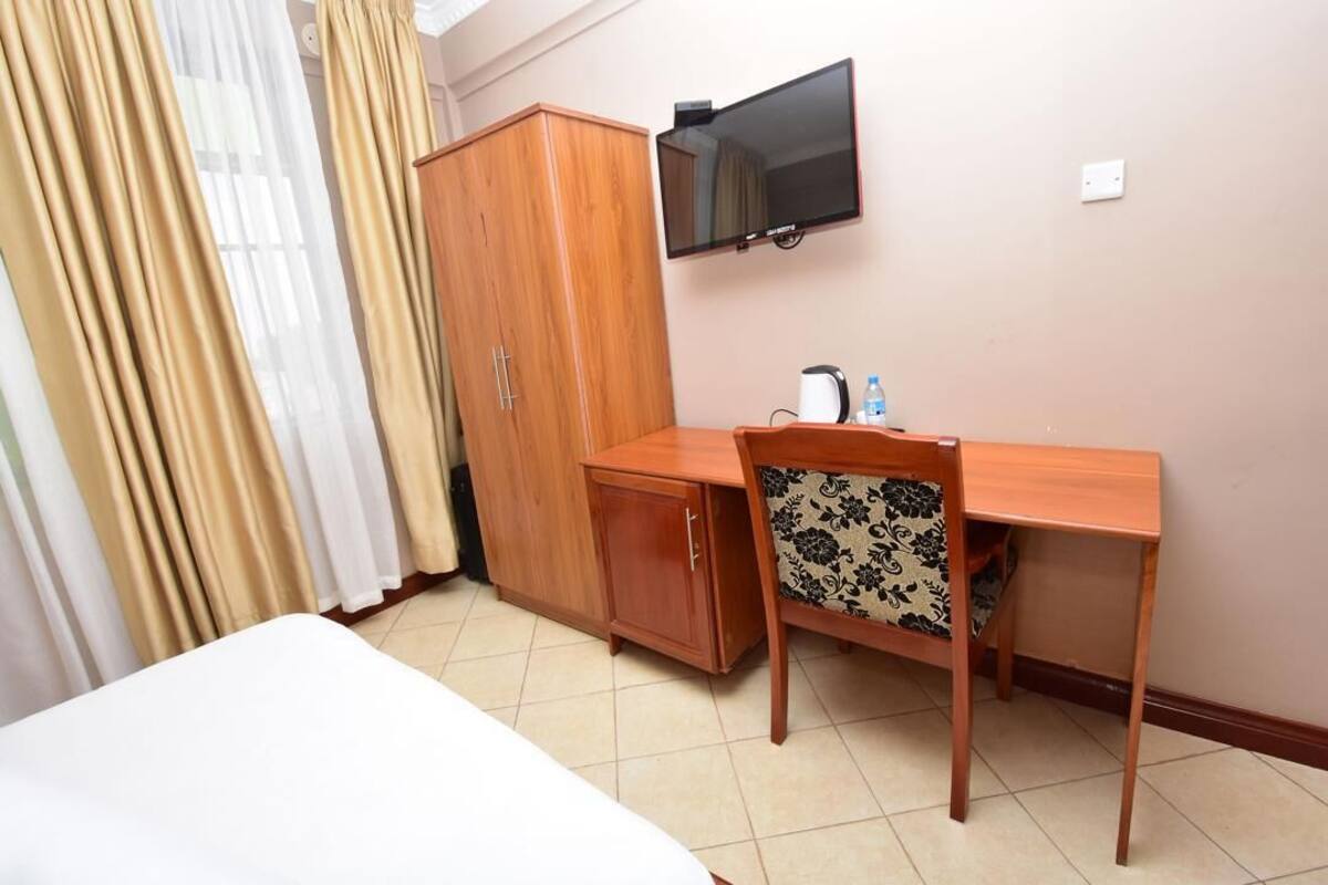 Une chambre d’hôtel comprenant un lit, un bureau en bois avec une chaise, une armoire, une télévision et une petite table avec une bouilloire et une bouteille d’eau.