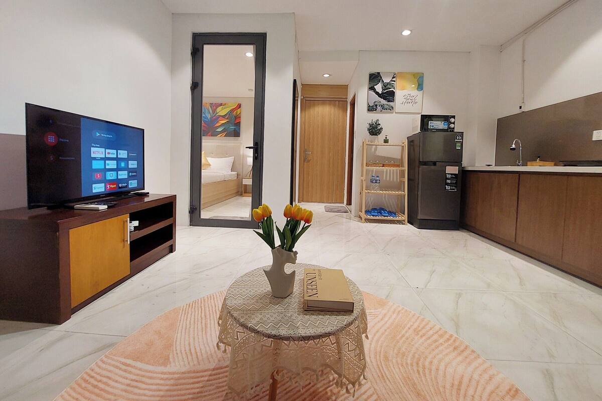 Una sala moderna con televisor de pantalla plana, una mesa de centro con un libro y flores, una zona de cocina con refrigerador y microondas, y un dormitorio visible a través de una puerta abierta.