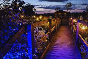 Eine Holzbrücke mit Geländern, umgeben von Pflanzen und einem pavillonartigen Bau im Hintergrund.
