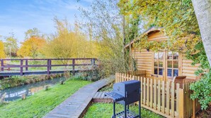 A wooden deck leading to a log cabin with a grill, surrounded by a wooden fence and a small pond.