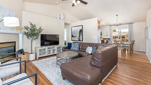 Un salón espacioso con un sofá de cuero marrón, una mesa de centro redonda y un televisor de pantalla plana. Hay una zona de comedor con sillas de madera y un candelabro.