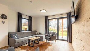 Una sala de estar moderna con un sofá gris, una mesa de centro de madera y un sillón gris. Hay un televisor de pantalla plana montado en la pared y un ventanal con cortinas.