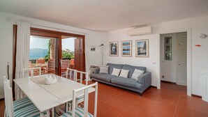 A living room with a sofa, a dining table with chairs, and a view of the sea through a glass door.