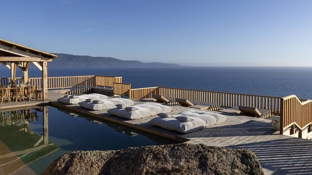A wooden deck with lounge chairs and pillows, overlooking a body of water and distant land.