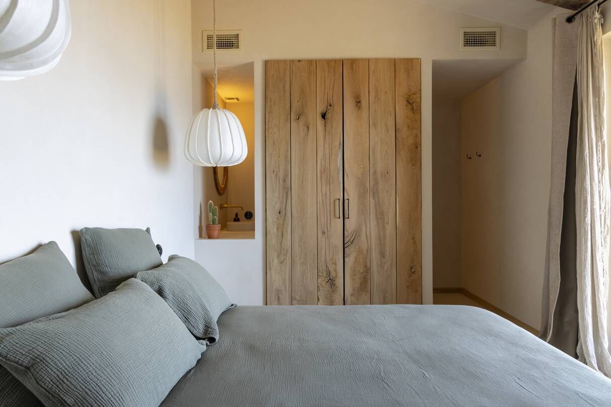 Un dormitorio con una cama, un armario de madera, una lámpara colgante y una pequeña estantería con una planta.