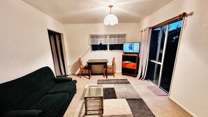 A living room with a dark green sofa, a small coffee table, a dining table with chairs, a TV on a wooden stand, and a large window with curtains.