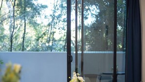 A dining area with a wooden table set for two, a white coffee table with a vase of flowers, and a sofa in the background.