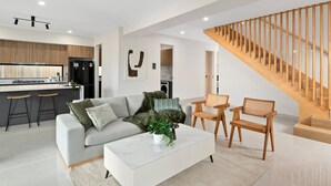 A modern living room with a sofa, coffee table, and chairs, a kitchen in the background, and a wooden staircase.