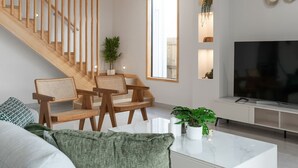 A modern living room with a wooden staircase, a large flat-screen TV, and a dining area with wooden chairs and a white table.
