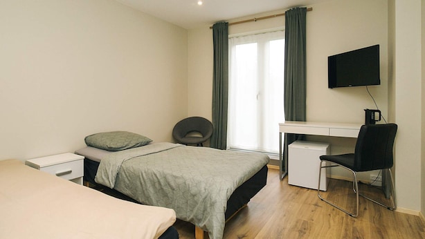 Quarto com cama, escrivaninha, cadeira, televisão e janela com cortinas.