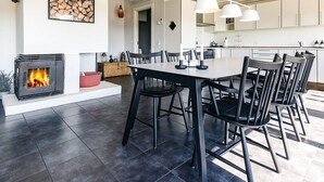 A modern kitchen with white cabinets, a dining area with a large table and chairs, and a fireplace with a visible fire. - 4 Person Holiday Park Home in Karrebaeksminde (Karrebæksminde)