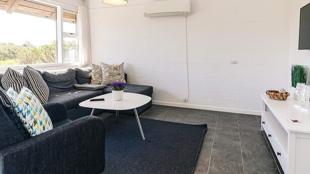 A modern living room with a grey sofa, patterned cushions, a round coffee table, and a white wall with an air conditioning unit. - 4 Person Holiday Park Home in Karrebaeksminde (Karrebæksminde)