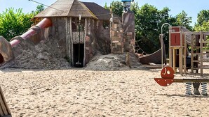 Une aire de jeux sablonneuse avec un petit bâtiment en pierre, une statue de Viking et un toboggan.