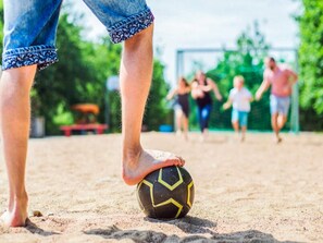 Iemand trapt een voetbal op een zandige ondergrond, met een doel in de verte.