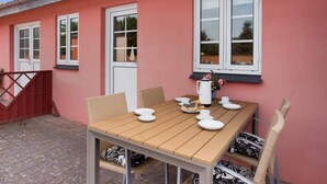 A red building with a wooden table set for dining, wicker chairs, and a window with a view of trees.