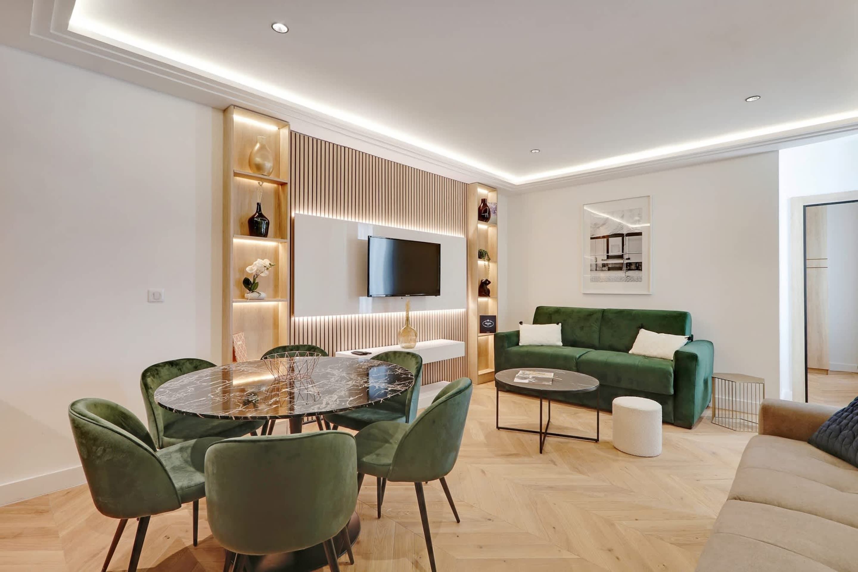 A modern living room with a round glass table, green upholstered chairs, a green sofa, and a flat-screen TV mounted on a wooden panel.