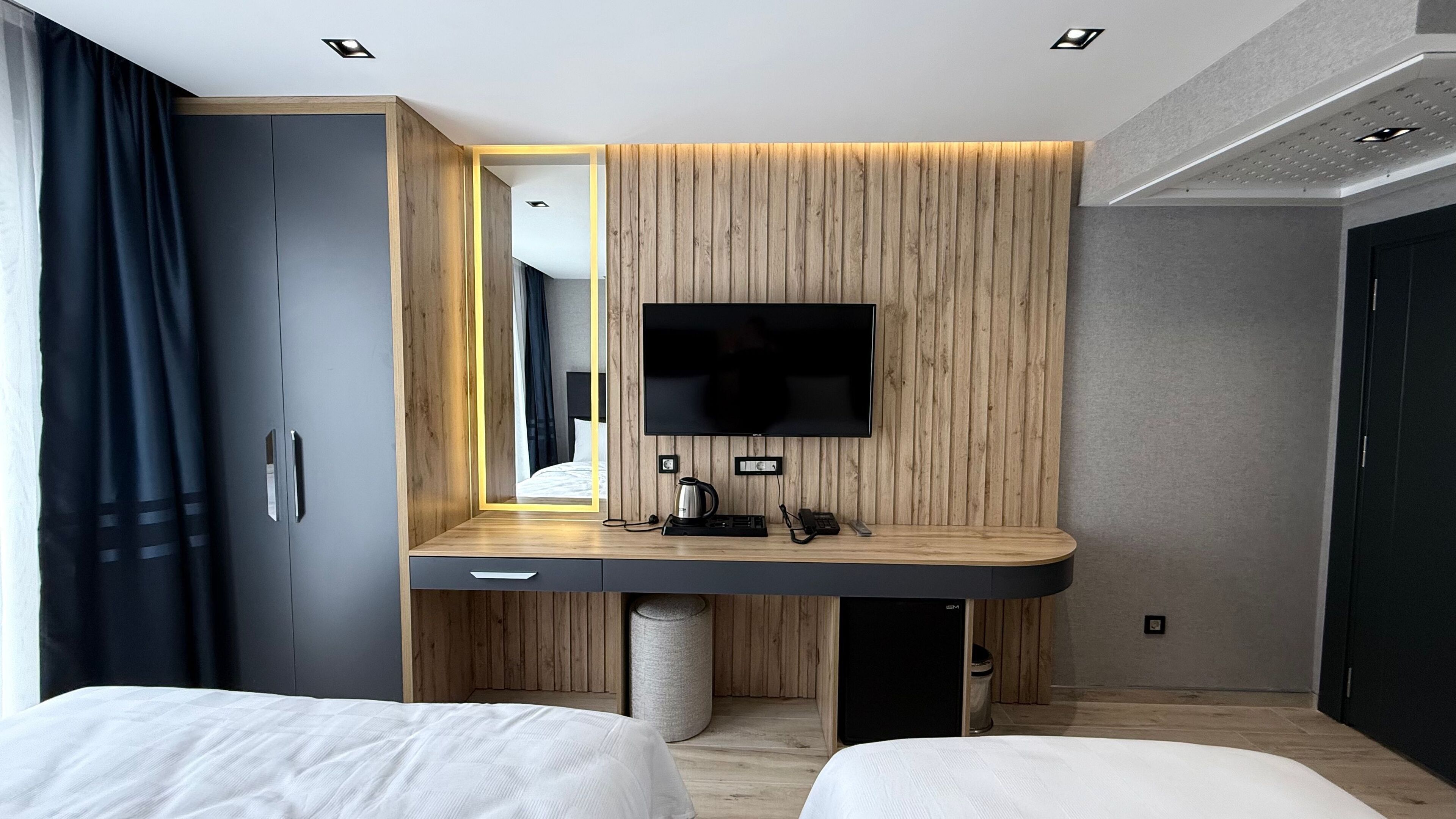 A modern hotel room with a wooden accent wall, a flat-screen TV mounted above a desk, a bed, and a large mirror.