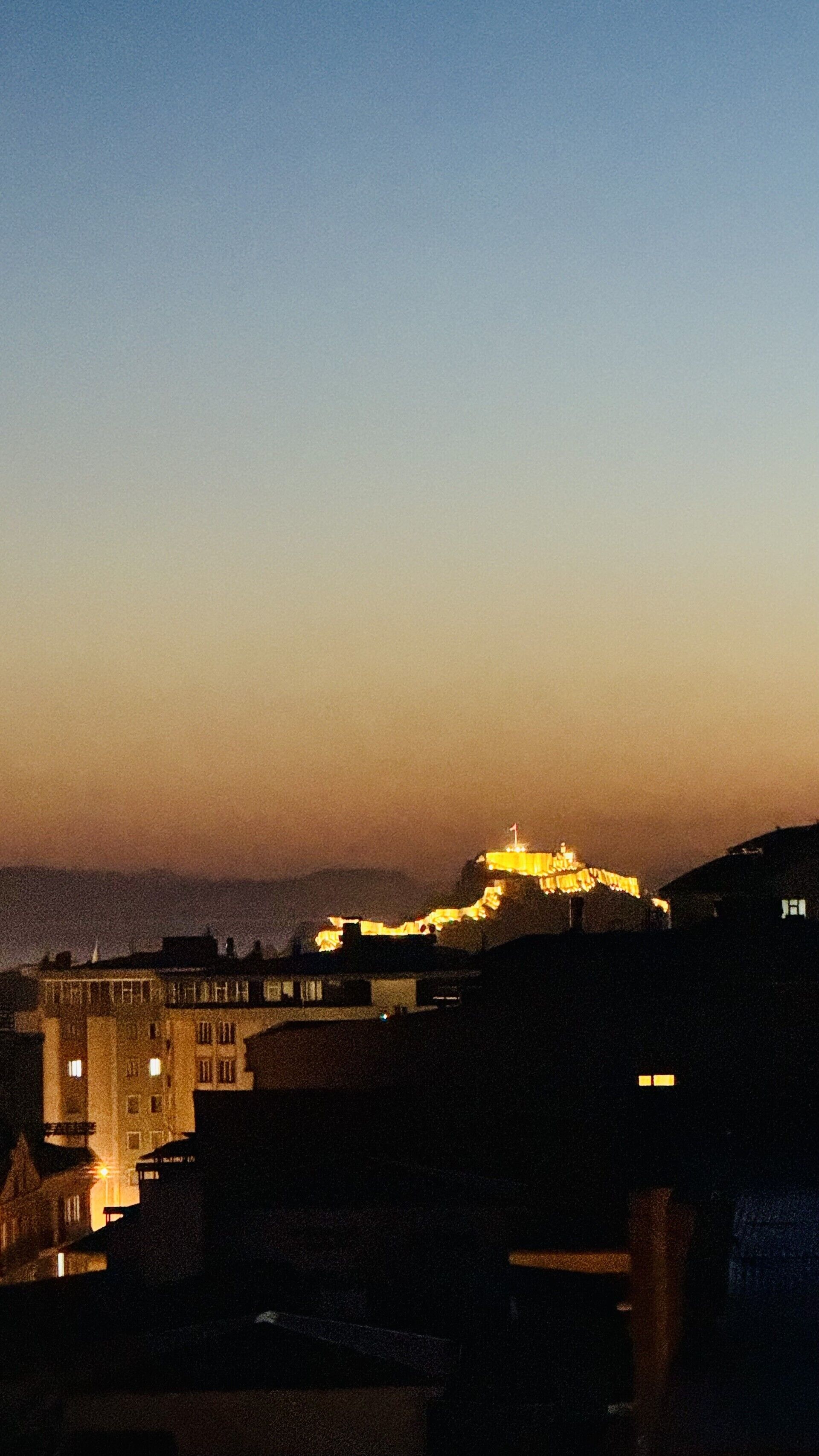 Vista noturna de um horizonte urbano com edifícios iluminados e uma estrutura imponente no topo de uma colina.