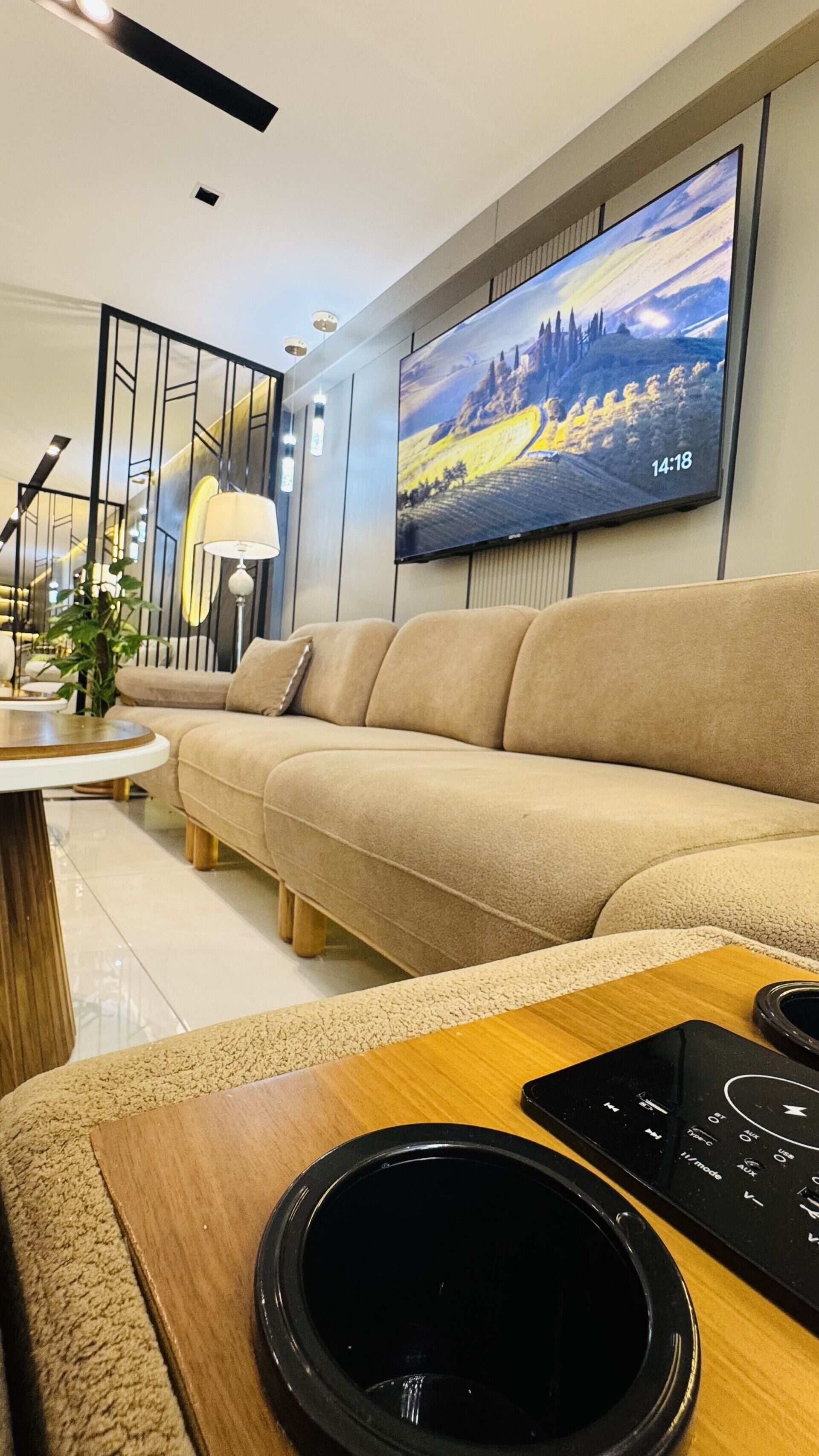 A modern living room with a beige sofa, a flat-screen TV mounted on the wall, a wooden coffee table, and a black speaker.