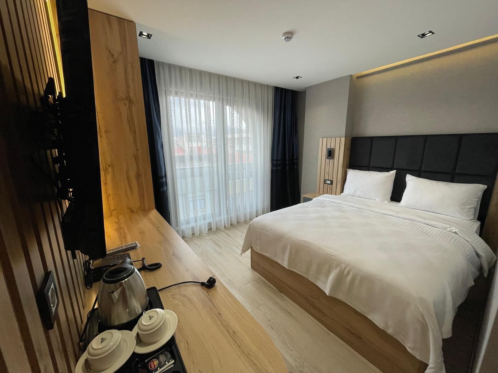 A modern hotel room with a large bed, a coffee maker, and a view of the city through sheer curtains.