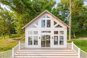 A white, two-story house with a wooden deck and white railings, surrounded by lush greenery. - Keenan Hollow Farm | 5B/4.5BA on River • Sleeps 21 (Niles)