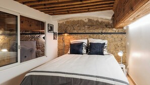 A bedroom with a stone wall, wooden ceiling beams, a bed with a striped bedspread, and a bedside lamp.