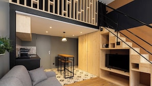 Ein modernes Wohnzimmer mit grauem Sofa, Holzboden, einem kleinen Tisch und einem Fernseher auf einem Holzständer.