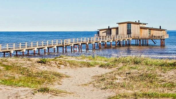 A wooden pier leading to a single-story wooden building with a flat roof, situated on a sandy beach with grass in the foreground. - 2 Person Holiday Park Home in Bastad (Bastad)