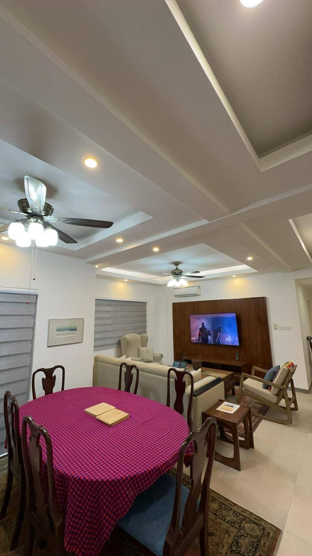 A modern living room with a dining area, ceiling fans, recessed lighting, and a flat-screen TV.
