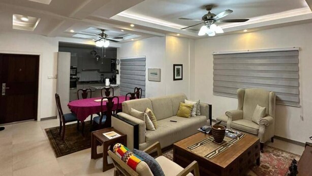 Una sala moderna con sofá, sillones y una mesa de centro. Hay una zona de comedor con una mesa y sillas rosas. La habitación tiene iluminación empotrada y un ventilador de techo.