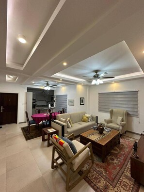 A modern living room with a sofa, armchairs, and a coffee table. There is a dining area with a pink table and chairs. The room has recessed lighting and a ceiling fan.