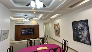 Un comedor con una mesa redonda cubierta por un mantel a cuadros, rodeada de sillas de madera. Hay un ventilador de techo sobre la mesa, y un televisor está montado en la pared al fondo.