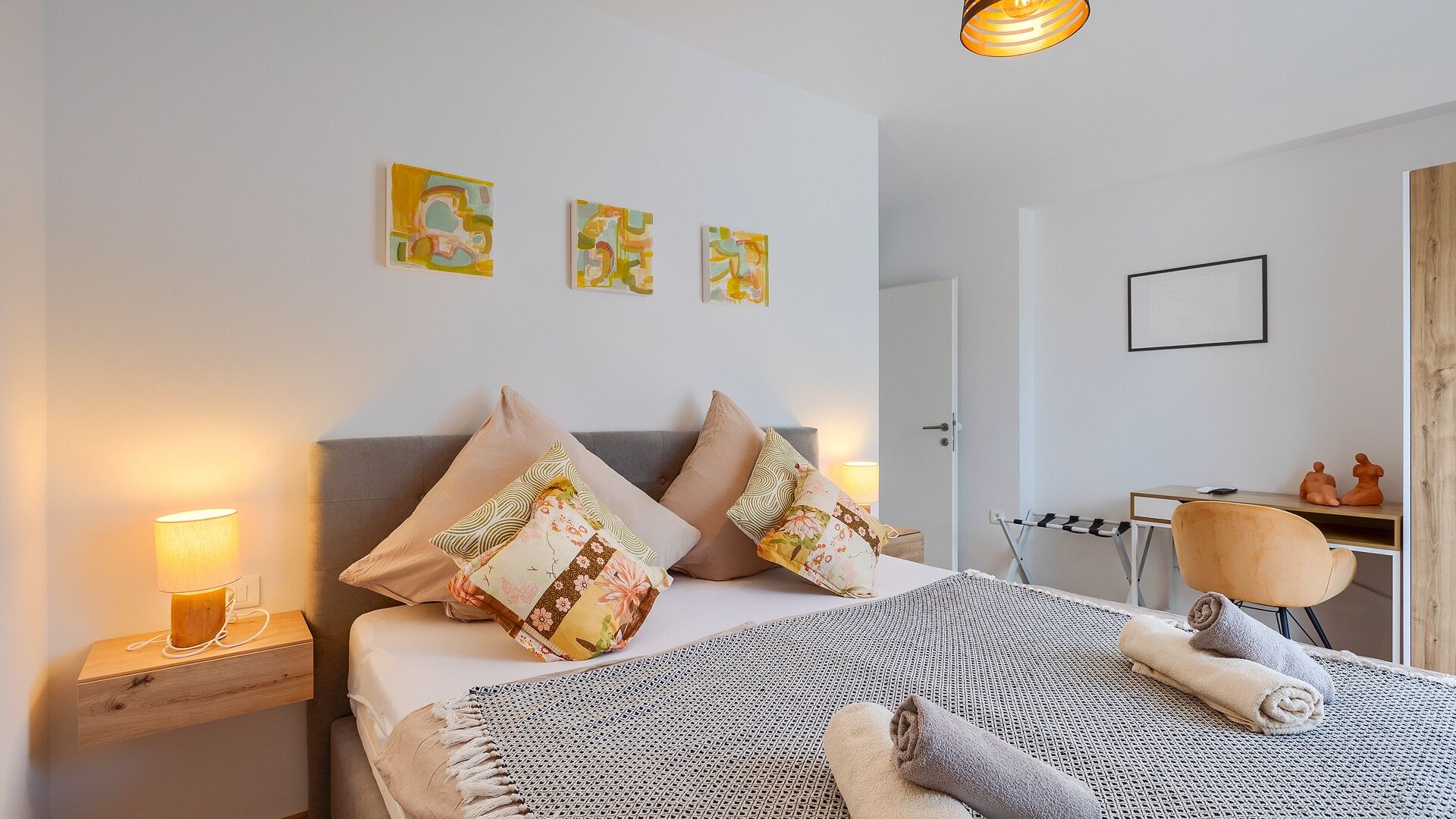 A neatly arranged bedroom with a bed, pillows, a bedside lamp, a desk, and a chair.