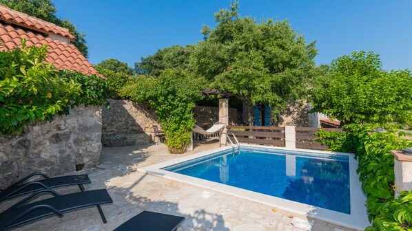 A swimming pool with stone tiles, surrounded by greenery and a stone wall.