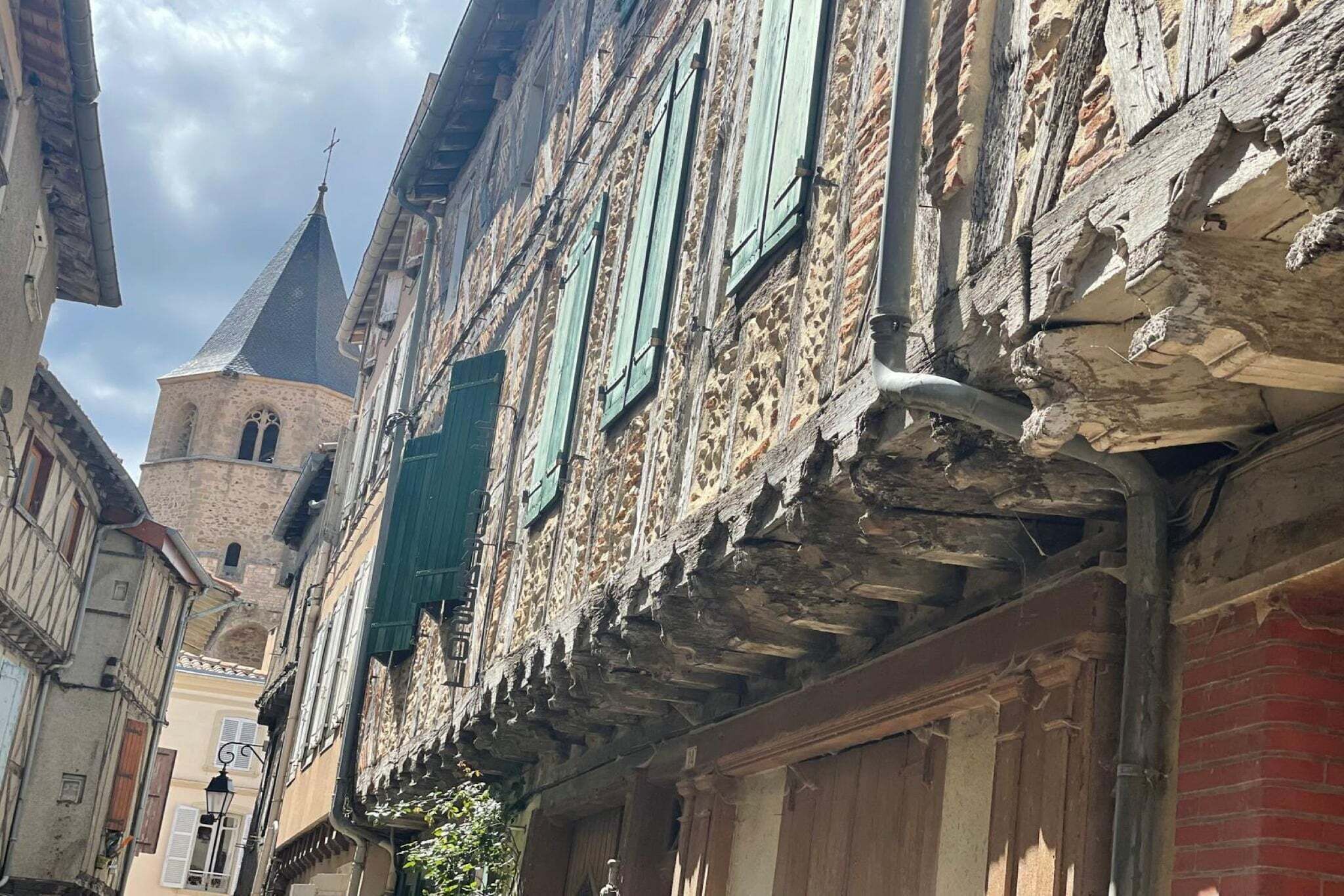 A narrow street with historic stone buildings, a church tower, and traditional shutters.