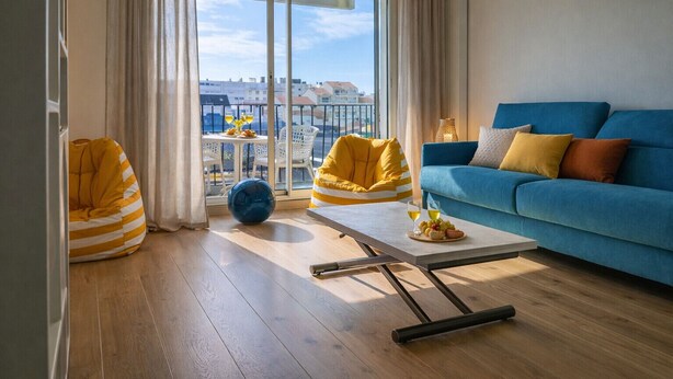 A modern living room with a blue sofa, yellow bean bag chairs, a coffee table with fruit and drinks, and a large window with a city view. - Charming apartment in the Presidents' District (Les Sables-d´Olonne)