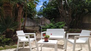 A patio area with white plastic chairs and a table, surrounded by greenery and a stone wall.