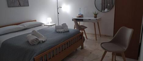 A bedroom with a bed, a desk, a chair, a lamp, a mirror, and a wall art piece.